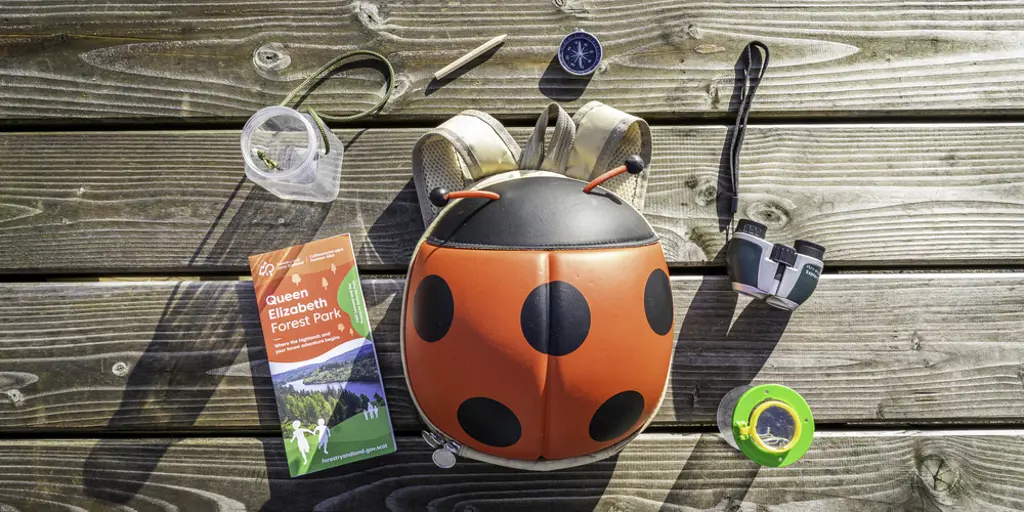 A red ladybird backpack on a table with a brochure, magnifying glass, compass and binoculars around it. 
