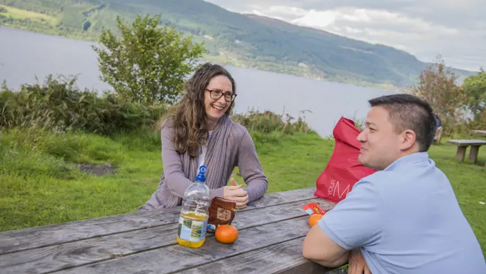 Two people at a picnic bench with Loch Ness in the background.