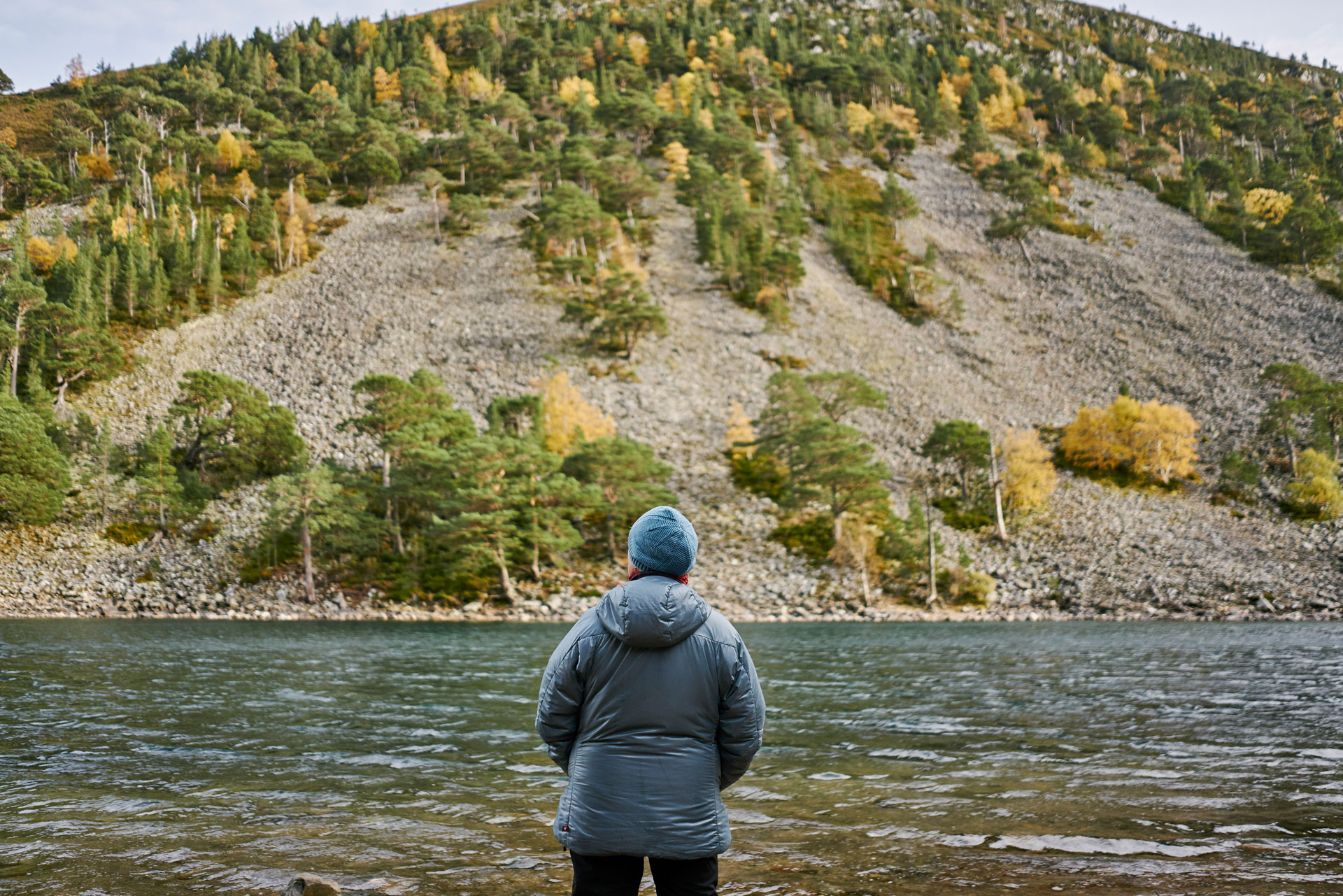 A person in a coat and hat looks over a body of water. On the other side is a steep bank with patchy tree cover. 
