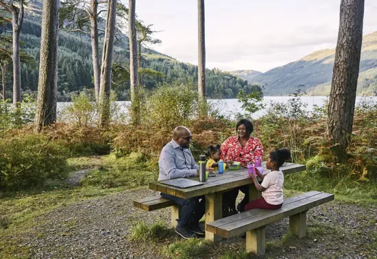  Man, woman and two young girls sit at picnic table with food and drink, at Jubilee Point, on shore of Loch Eck