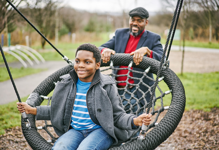 Young boy sits on the basket swing pushed by father in play area at Cuningar Loop Woodland Park, Glasgow