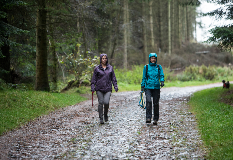 Two women in rain jackets holding dog leashes with a dog walking behind them