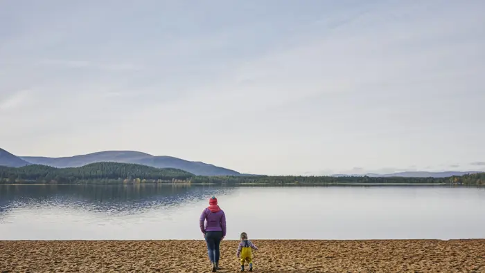 Young woman with purple jacket walks with toddler, on sand at Loch Morlich beach, Glenmore Forest Park, near Aviemore