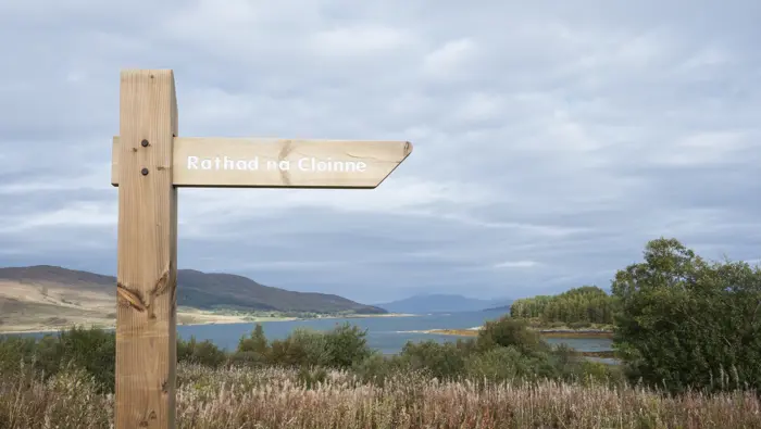 Wooden sign reading Rathad na Cloinne at Broadford forest on the Isle of Skye. Wild plants, flowers and trees in the background with a view towards the sea and distant hills.