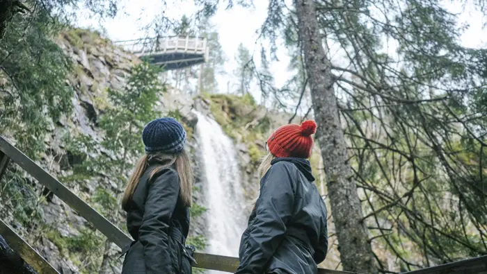 Two young women, one with orange woollen hat, other with blue striped woollen hat, look up from wooden viewing platform to high point of Plodda Falls waterfall as it roars down rockface, Glen Affric