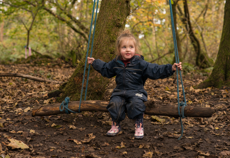 Young girl in blue wet weather gear sits on swing made of log, in Rannoch Wood, Renfrewshire Woods near Johnstone
