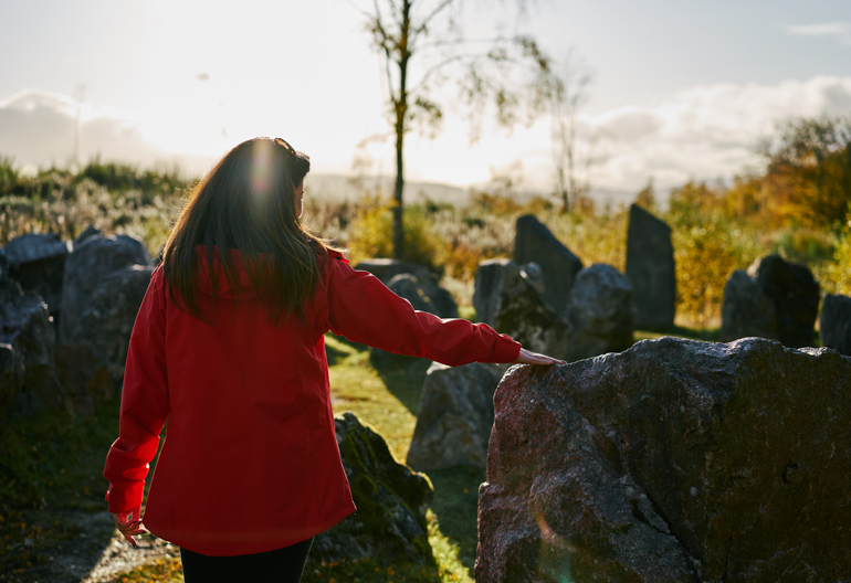 Rear view of woman in evening sun, wearing red jacket, touching boulders in Touchstone Maze, labyrinth of Scottish stones, Blackmuir Wood, near Strathpeffer