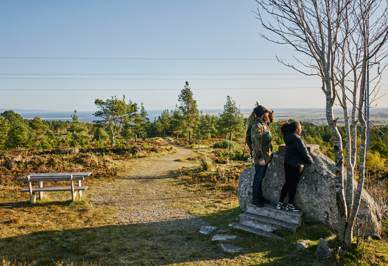 Man and teenage girl stand on open ground at Pulpit Rock, Tain Hill looking at panoramic views over Tarbet Ness