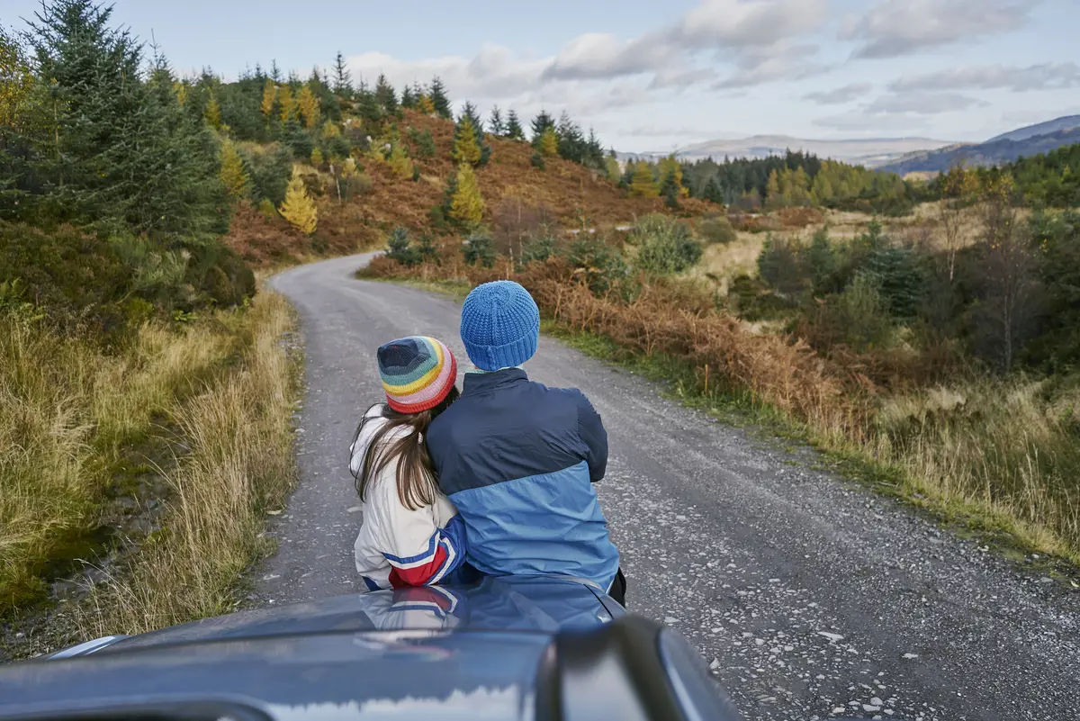 Two people leaning on car bonnet looking towards forest