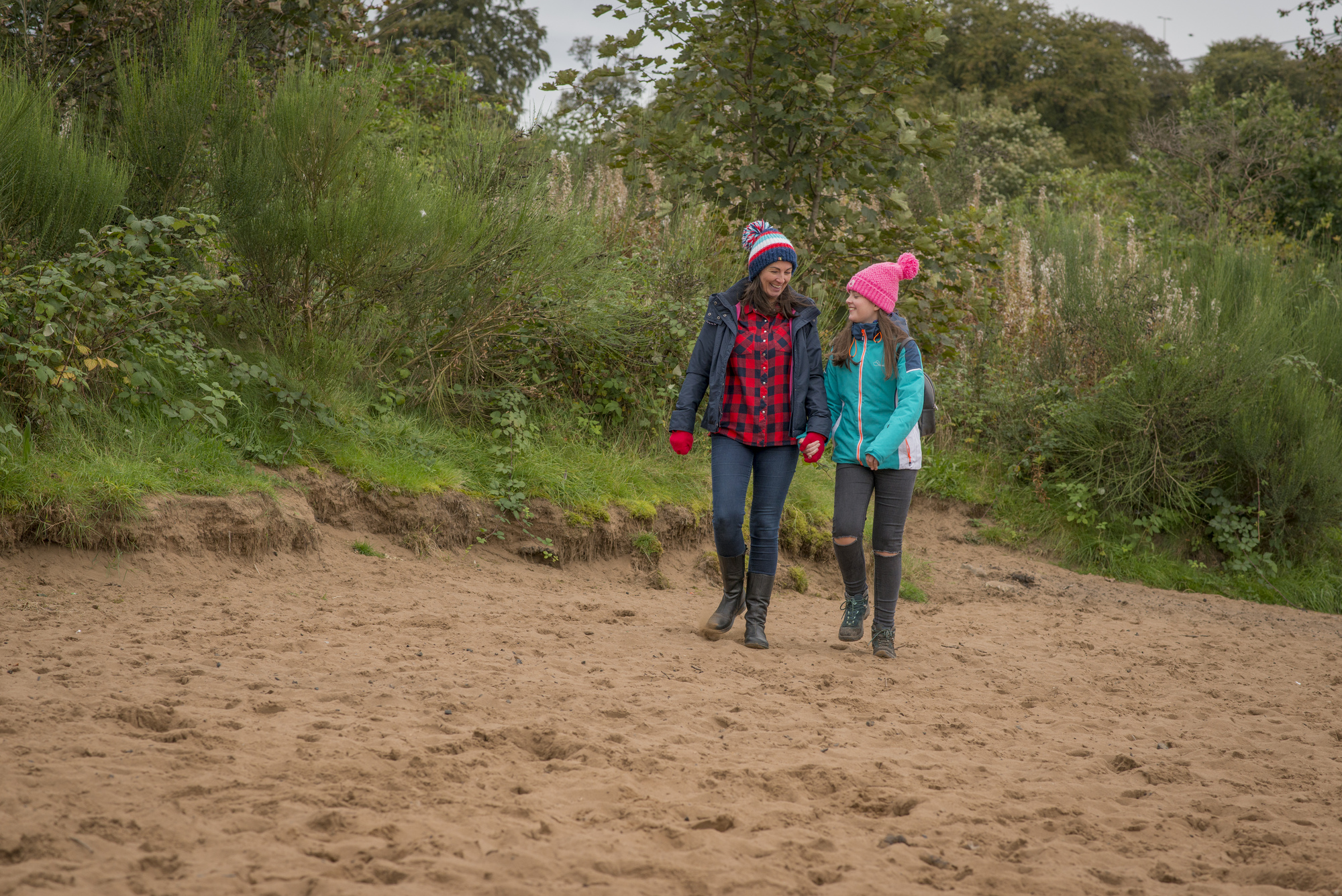  Woman and young girl walk hand in hand along the sand with woodland in background, at Boden Boo riverside beach, Renfrewshire Woods, near Erskine Bridge