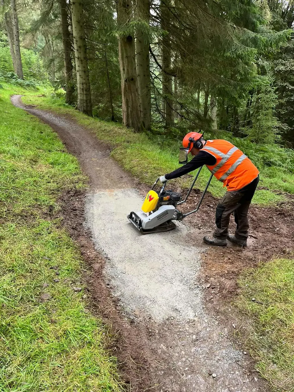 A person in hi-vis and hard hat uses a wacker machine to compress the repaired trail.