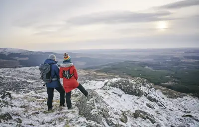 A couple standing on a snowy hillside with expansive views of forestry and farmland.