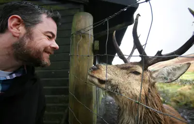 A male visitor stands close to a wire fence with a stag deer standing across from him