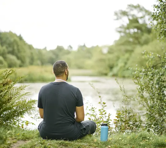 Man in black t shirt with blue flask beside him, sits on grass edge of Bordie Loch, Devilla Forest, near Kincardine