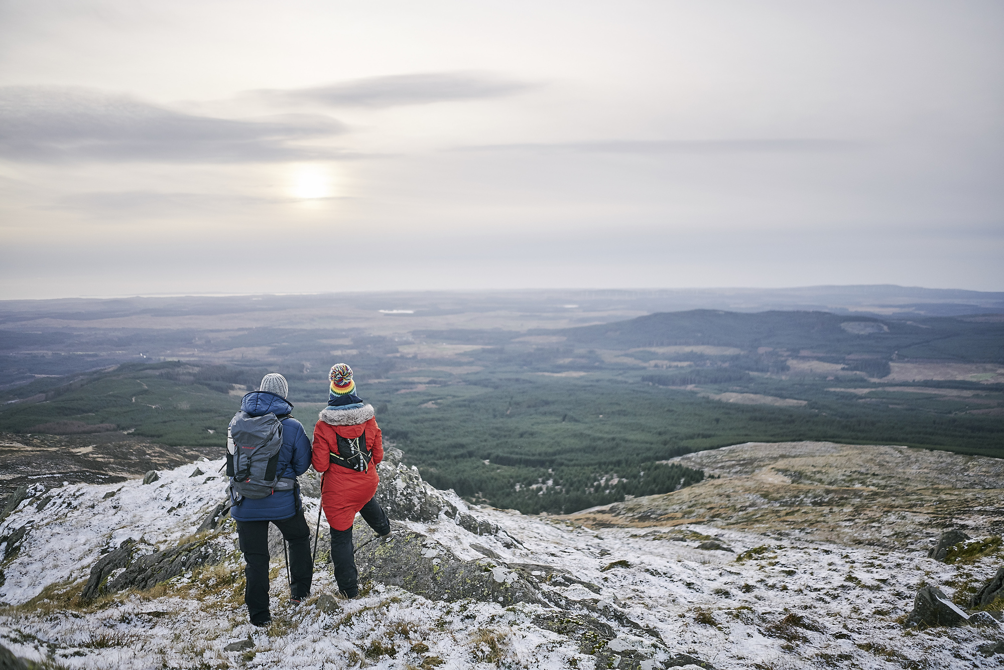 Male hiker in blue jacket and grey hat stands on snowy ground with female hiker in red jacket and rainbow pompom hat, both carrying rucksacks, looking over panoramaic view from Bennan Hill, Glentrool to plain below.
