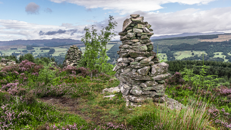 Stone carin built on the top of a hill with heather growing and a valley beyond with trees and farms