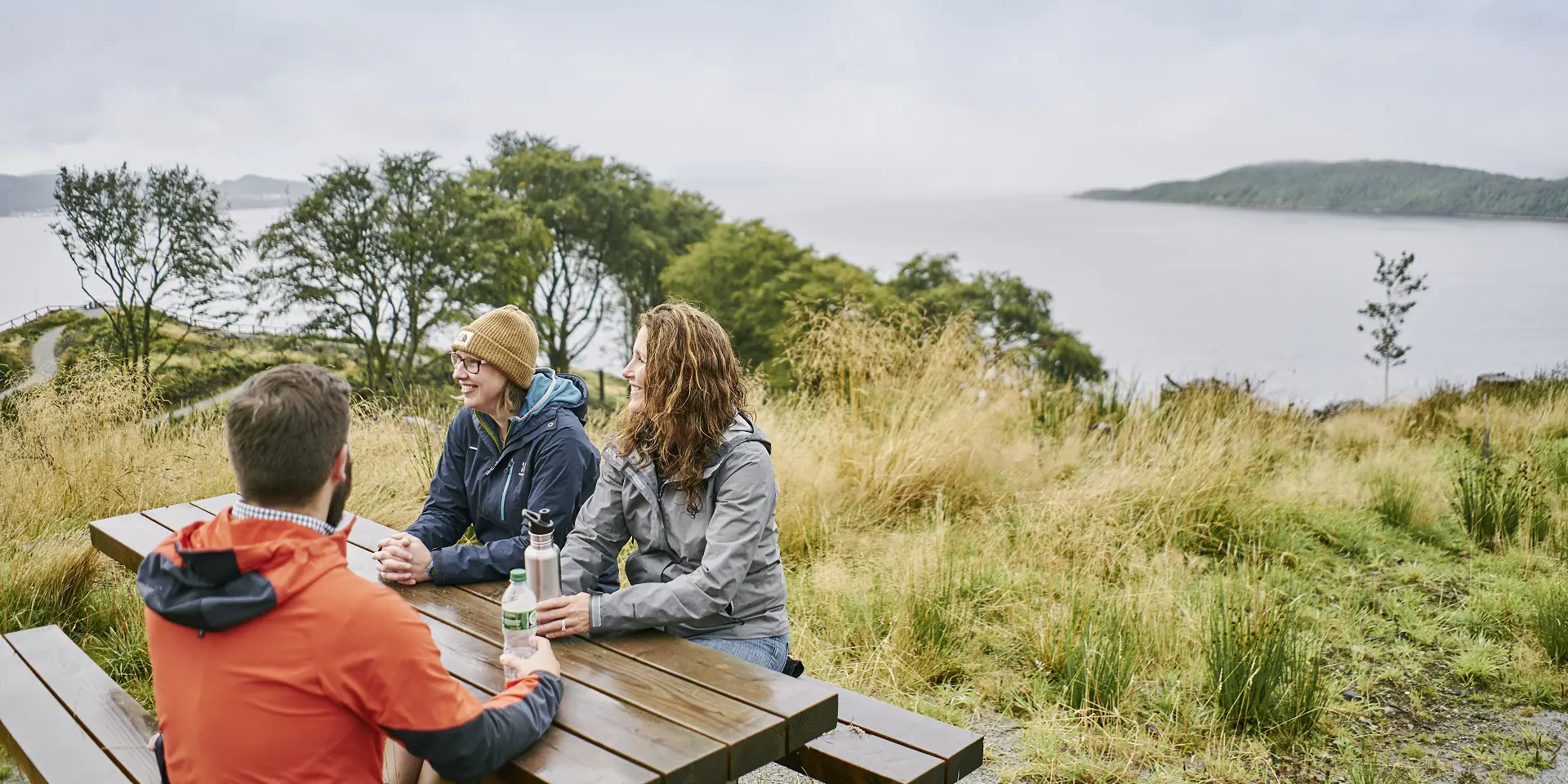 Two women and a man sitting at a picnic bench overlooking trees and sea