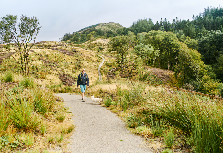 Young woman with small dog walking on coastal trail with forest in background, near Beinn Lora, Benderloch