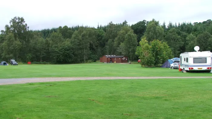 Green grassy field with gravel road through it and trees beyond, with a parked caravan