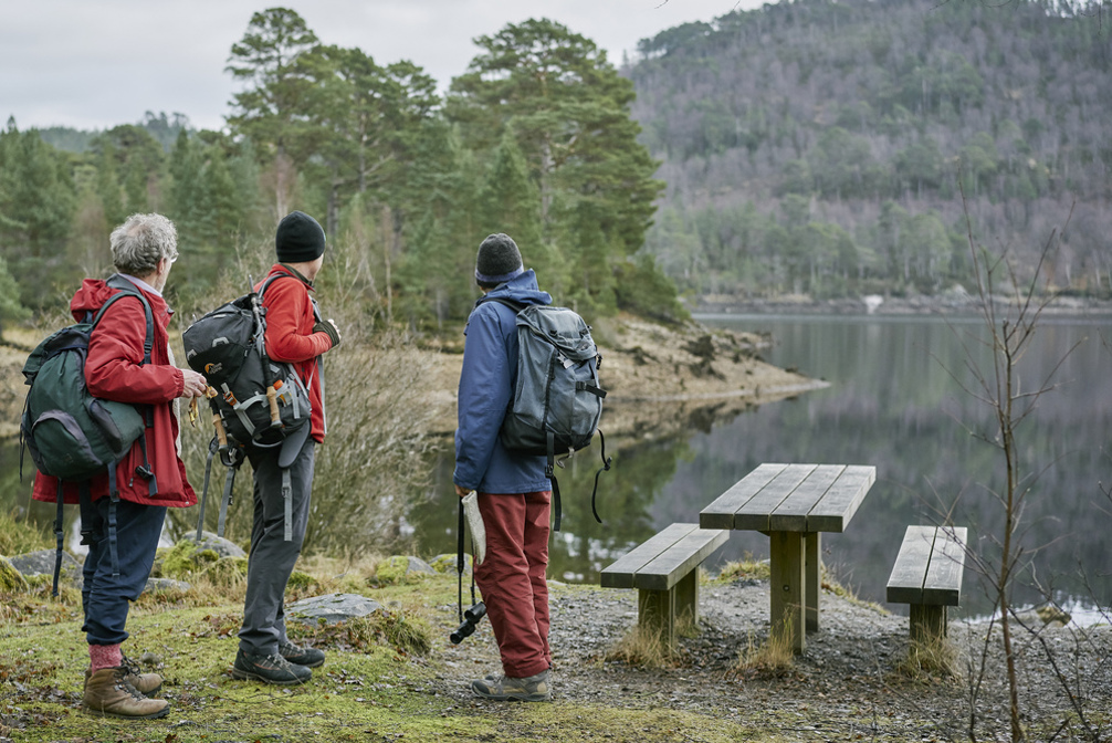 Tree men standing next to a loch and picnic table surrounded by forest