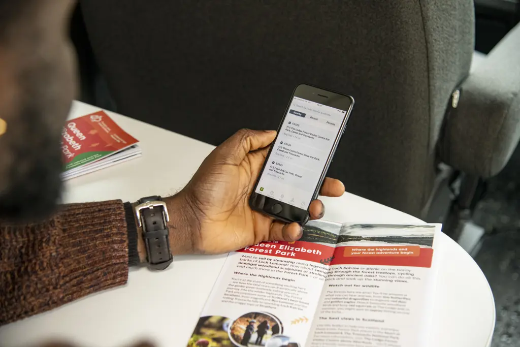 Man's hand holding a mobile phone on which the RingGo app is opened, above a motorhome/campervan table on which a leaflet sits