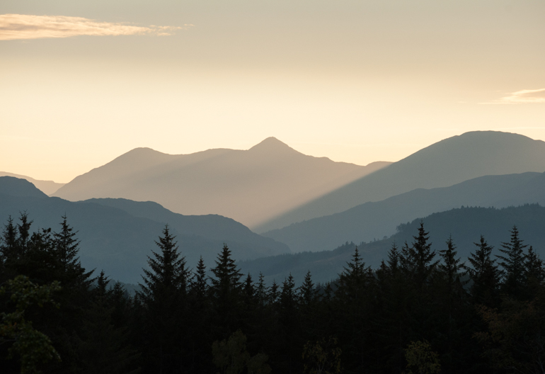 Silhouettes of distant hills, mountains and conifer forest beneath a sunny and cloudy evening sky from Blackmuir Wood
