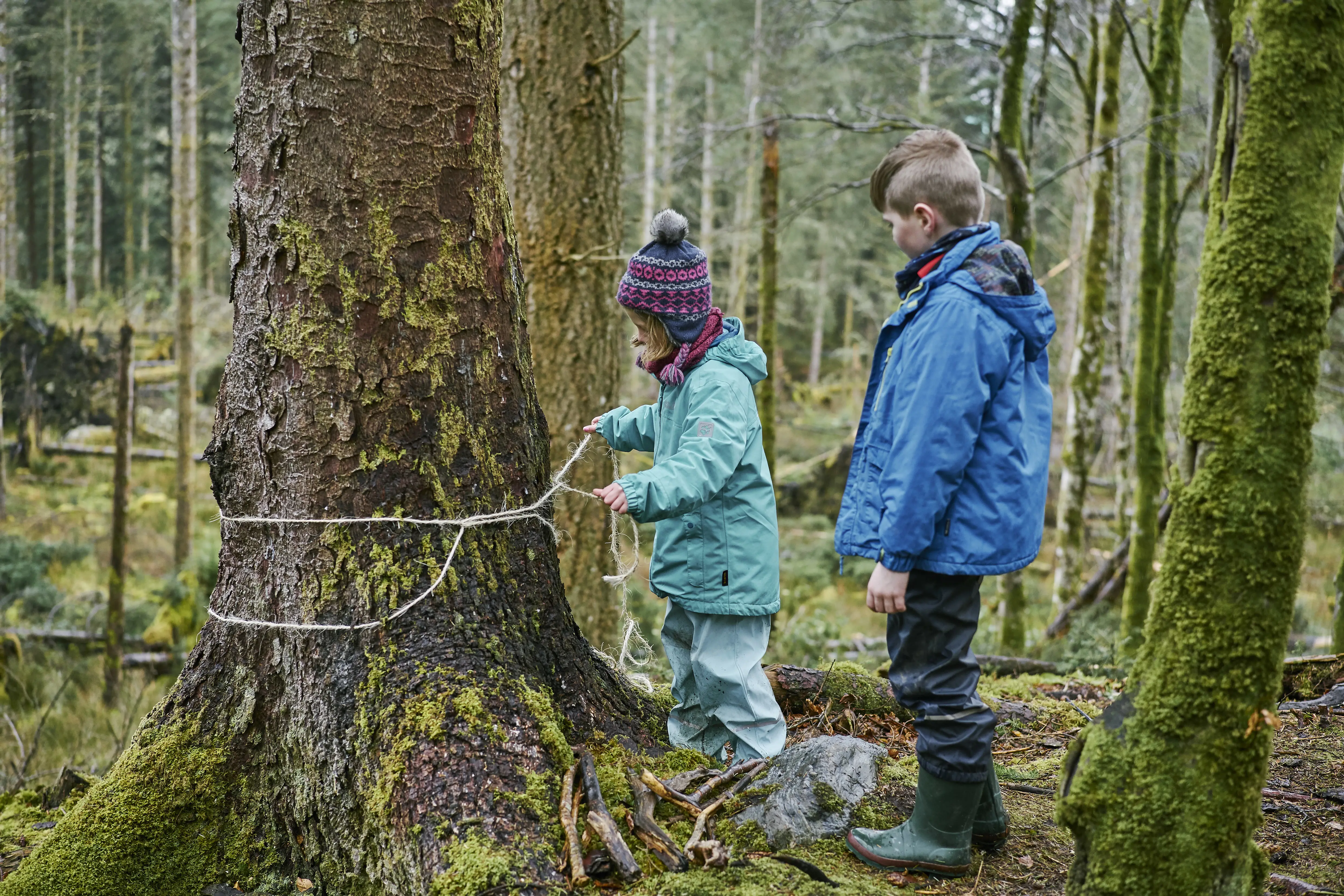 Young girl in turquoise wet weather gear and woollen hat ties string around trees, watched by young boy in blue wet weather clothes, at Sutherland's Grove, Barcaldine