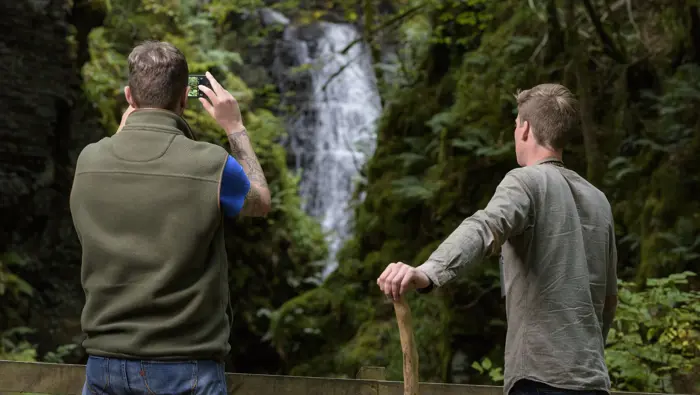 Two people looking at a waterfall with one taking a picture on their phone.