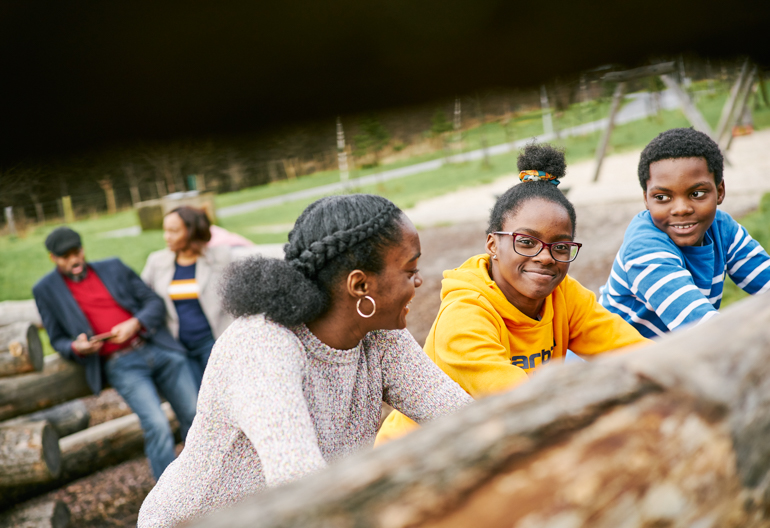 Two teenage girls and young boy climbing up logs at play area in Cuningar Loop Woodland Park, Glasgow (adult couple blurred in background)